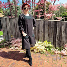 Load image into Gallery viewer, Sandy-tunic-dress-with-tab-button-long-sleeves-jewel-neckline-applique-detail-on-front-yoke-and-pockets-and-string-tie-waist-in-black-next-to-fence-and-pink-tree-2
