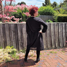 Load image into Gallery viewer, Sandy-tunic-dress-with-tab-button-long-sleeves-jewel-neckline-applique-detail-on-front-yoke-and-pockets-and-string-tie-waist-in-black-next-to-fence-and-pink-tree-back-view
