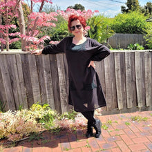 Load image into Gallery viewer, Sandy-tunic-dress-with-tab-button-long-sleeves-jewel-neckline-applique-detail-on-front-yoke-and-pockets-and-string-tie-waist-in-black-next-to-fence-and-pink-tree
