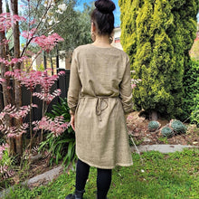 Load image into Gallery viewer, Sandy-tunic-dress-with-tab-button-long-sleeves-jewel-neckline-applique-detail-on-front-yoke-and-pockets-and-string-tie-waist-in-sand-next-to-pink-tree-back-view
