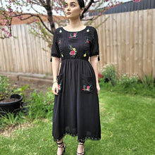 Load image into Gallery viewer, ashley-embroidered-midi-dress-with-embroidery-and-lace-detail-on-bodice-and-front-pockets-and-lace-on-hem-trim-in-black-front-of-tree
