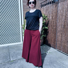 Load image into Gallery viewer, cotton-palazzo-pants-with-extra-wide-leg-fitted-mid-waist-elasticated-at-back-and-side-pockets-in-burgundy-worn-with-black-tshirt-front-of-gate
