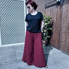 Load image into Gallery viewer, cotton-palazzo-pants-with-extra-wide-leg-fitted-mid-waist-elasticated-at-back-and-side-pockets-in-burgundy-worn-with-black-tshirt-front-of-gate
