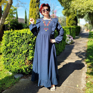 ginger-long-sleeve-embroidered-maxi-dress-with-ruffled-trim-over-shoulders-cinched-waistline-with-sash-tie-and-ruffled-hem-in-teal-next-to-garden-hedge