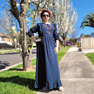 ginger-long-sleeve-embroidered-maxi-dress-with-ruffled-trim-over-shoulders-cinched-waistline-with-sash-tie-and-ruffled-hem-in-teal-next-to-trees-in-bloom