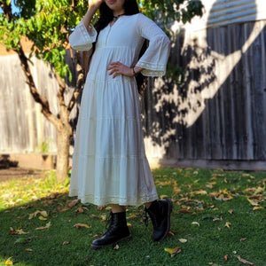 Sharon_tiered_midi_dress_in_cream_with_Vneckline_fitted_buttoned_bust_tiered_skirt_with_lace_trim_at_hem_and_bell_sleeves_with_lace_trim_with_boots_front_of_garden_tree